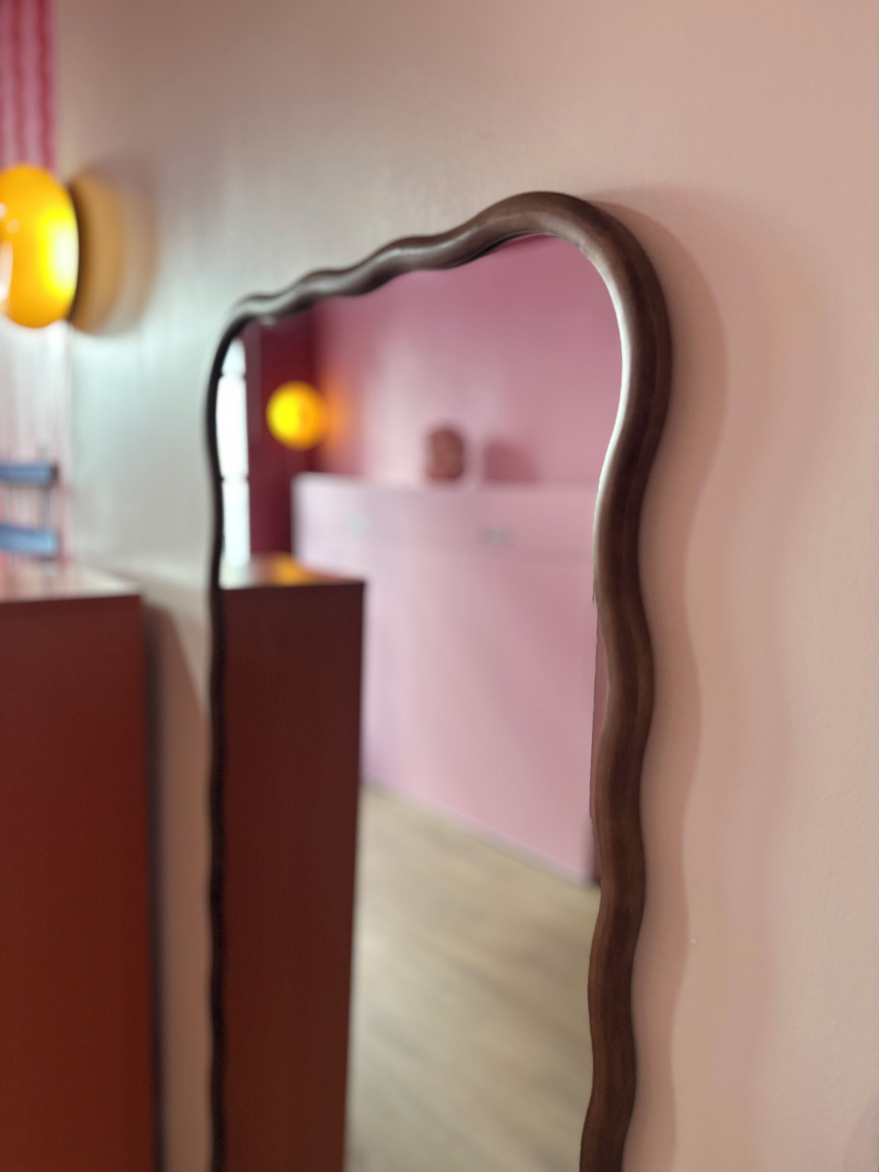 A mirror with a wavy frame reflecting a room with a pink wall and a bed.
