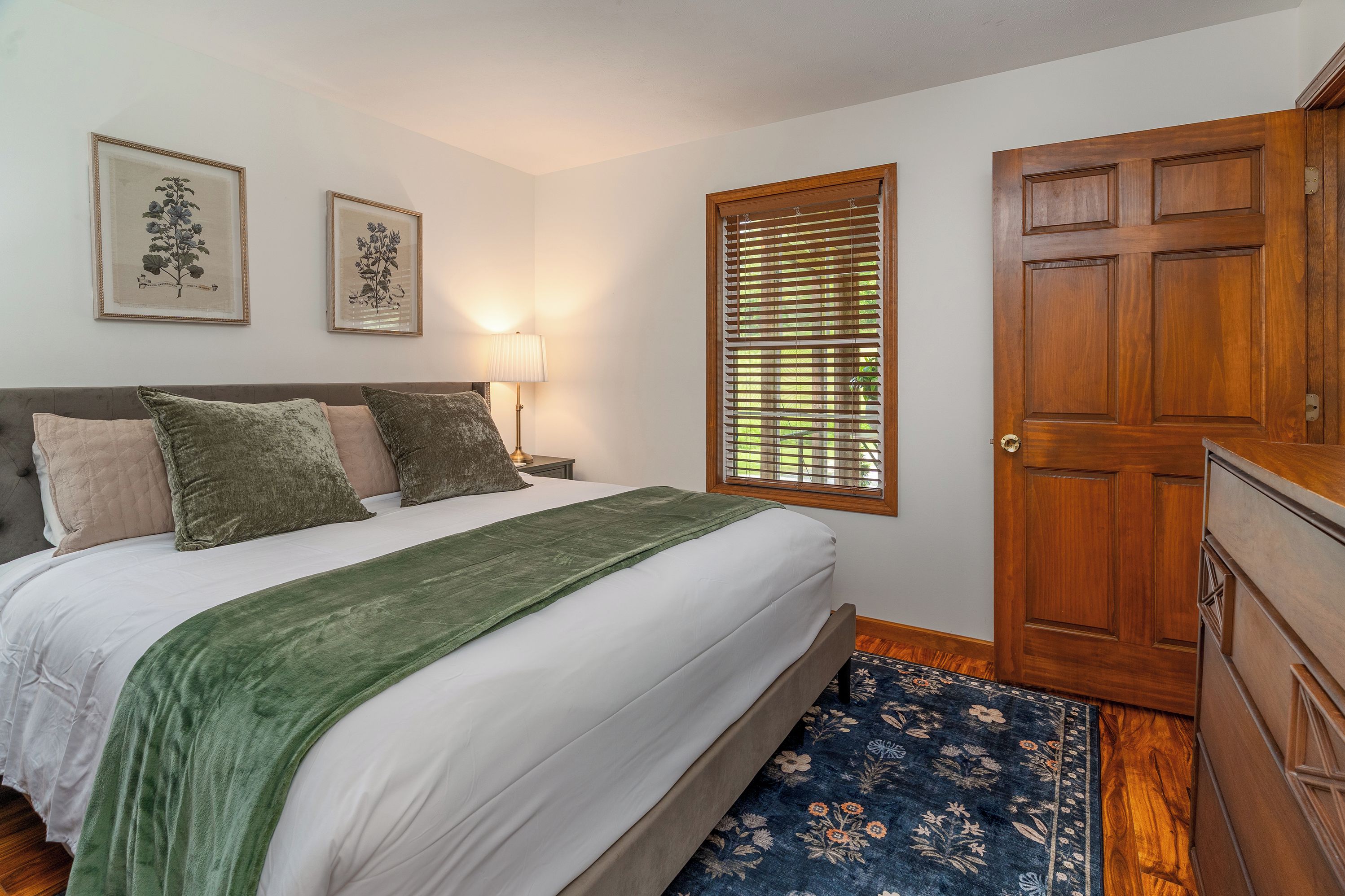 A bedroom with a bed, a nightstand, a lamp, a door, and two framed botanical prints on the wall.
