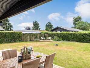 Eine Terrasse mit Tisch und Stühlen, ein Rasen und ein Heckenstreifen.