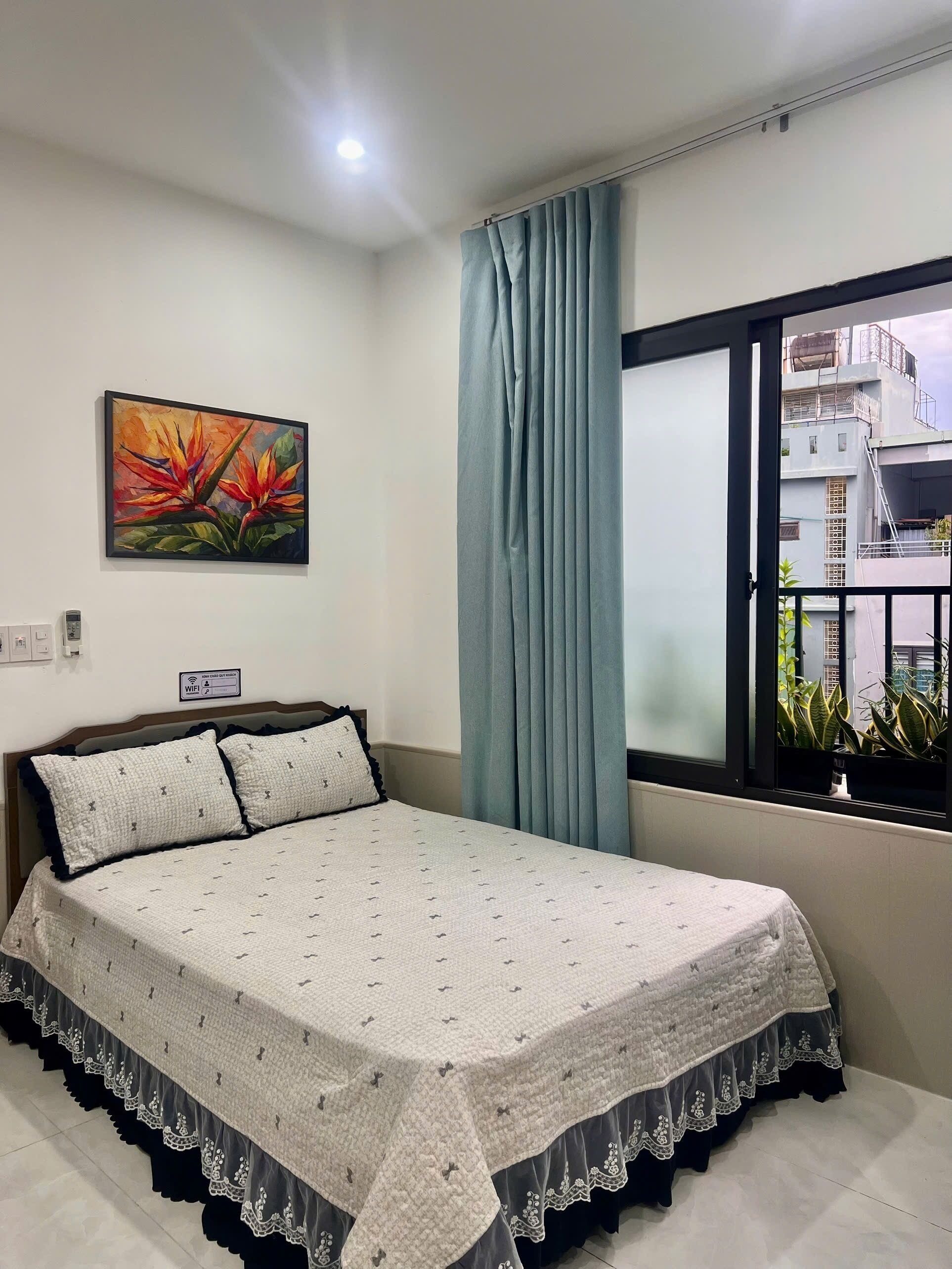 A hotel room with a bed, a painting, a window with curtains, and a view of buildings.