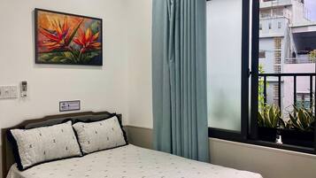 A hotel room with a bed, a painting, a window with curtains, and a view of buildings.