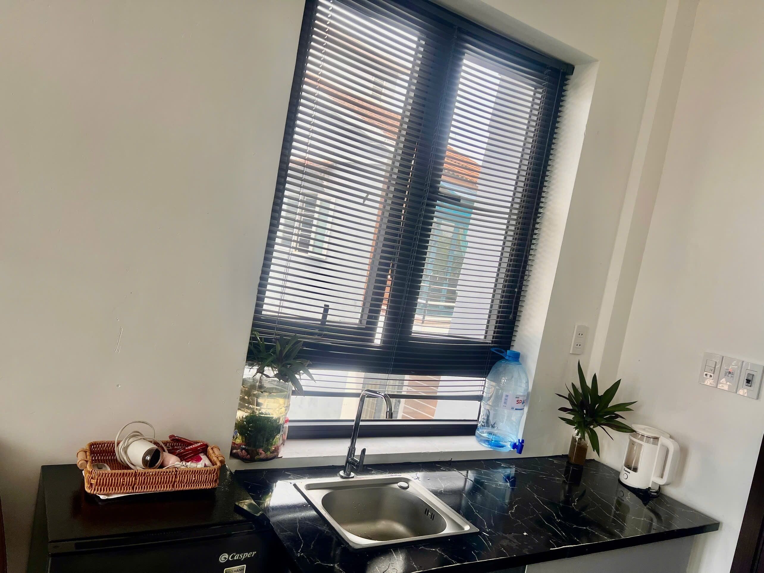 A kitchen with a black countertop, a sink, a window with blinds, a water dispenser, a kettle, and a basket with items.