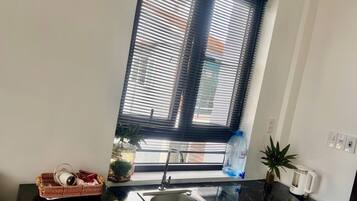 A kitchen with a black countertop, a sink, a window with blinds, a water dispenser, a kettle, and a basket with items.