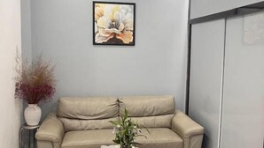 Een kamer met een beige bank, een ronde tafel met een vaas, en een bloemig kunstwerk in een lijst aan de muur.