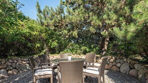 Une terrasse extérieure avec une table ronde et des chaises en osier, entourée d’un mur de pierre et d’une végétation luxuriante.