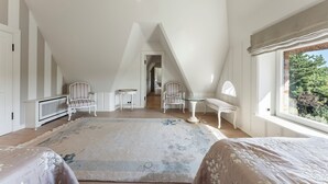 Une chambre spacieuse située dans les combles, avec un plafond haut, une grande fenêtre donnant sur les arbres et un coin salon confortable avec une table et des chaises.