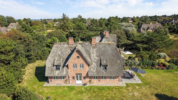 Une grande maison à toit de chaume, aux murs en briques, entourée d’une végétation luxuriante et d’une pelouse bien entretenue.