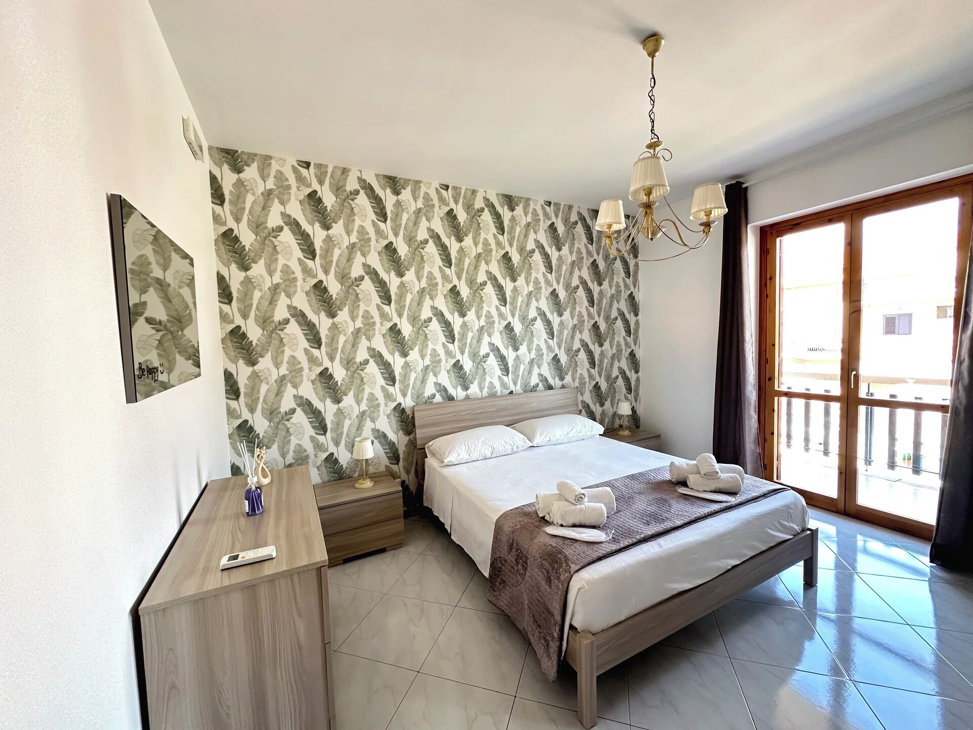 A hotel room with a bed, bedside tables, a nightstand, a mirror, a chandelier, and a view through a large window.