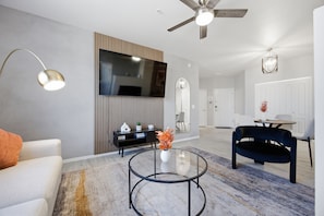 A modern living room with a glass coffee table, a sofa, a black chair, a ceiling fan, and a flat-screen TV mounted on the wall. - Location • Pool • Lux Gym • Free Parking • Cozy Retreat • Fireplace (Phoenix)