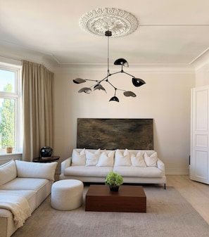 A modern living room with a white sofa, a round ottoman, a wooden coffee table, and a unique ceiling light fixture.