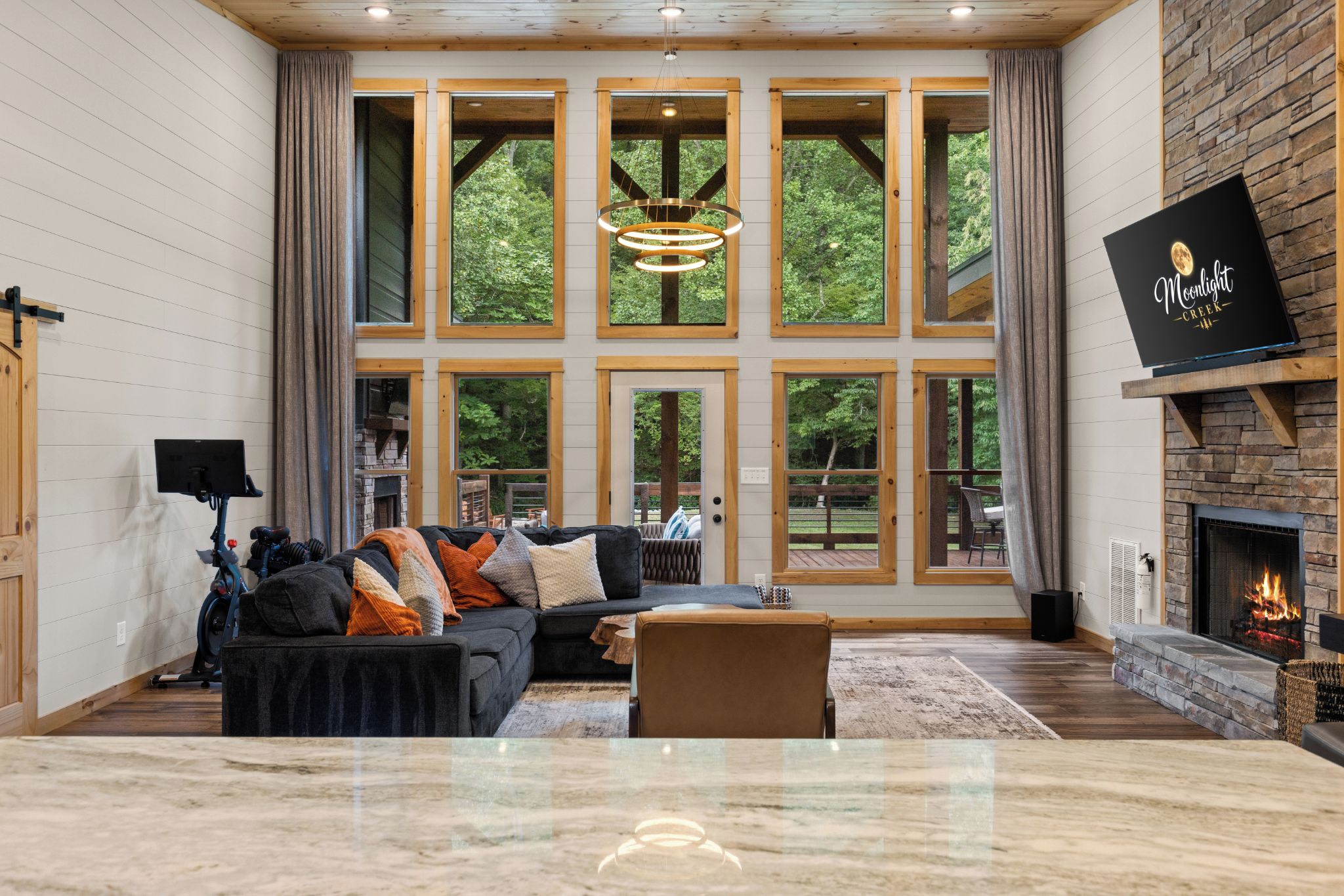 A spacious living room with a large fireplace, a stone accent wall, and a view of the outdoors through large windows.