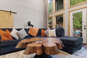 A modern living room with a dark grey sectional sofa, wooden coffee table, and a large window offering a view of greenery. - Private Creekfront Luxury: Sauna · Cold Plunge · Hot Tub · Fire pit · 4-acres (Mineral Bluff)