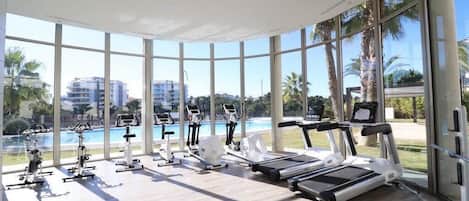 Un gimnasio con diversos equipos de ejercicio, amplias ventanas y vista a una piscina.