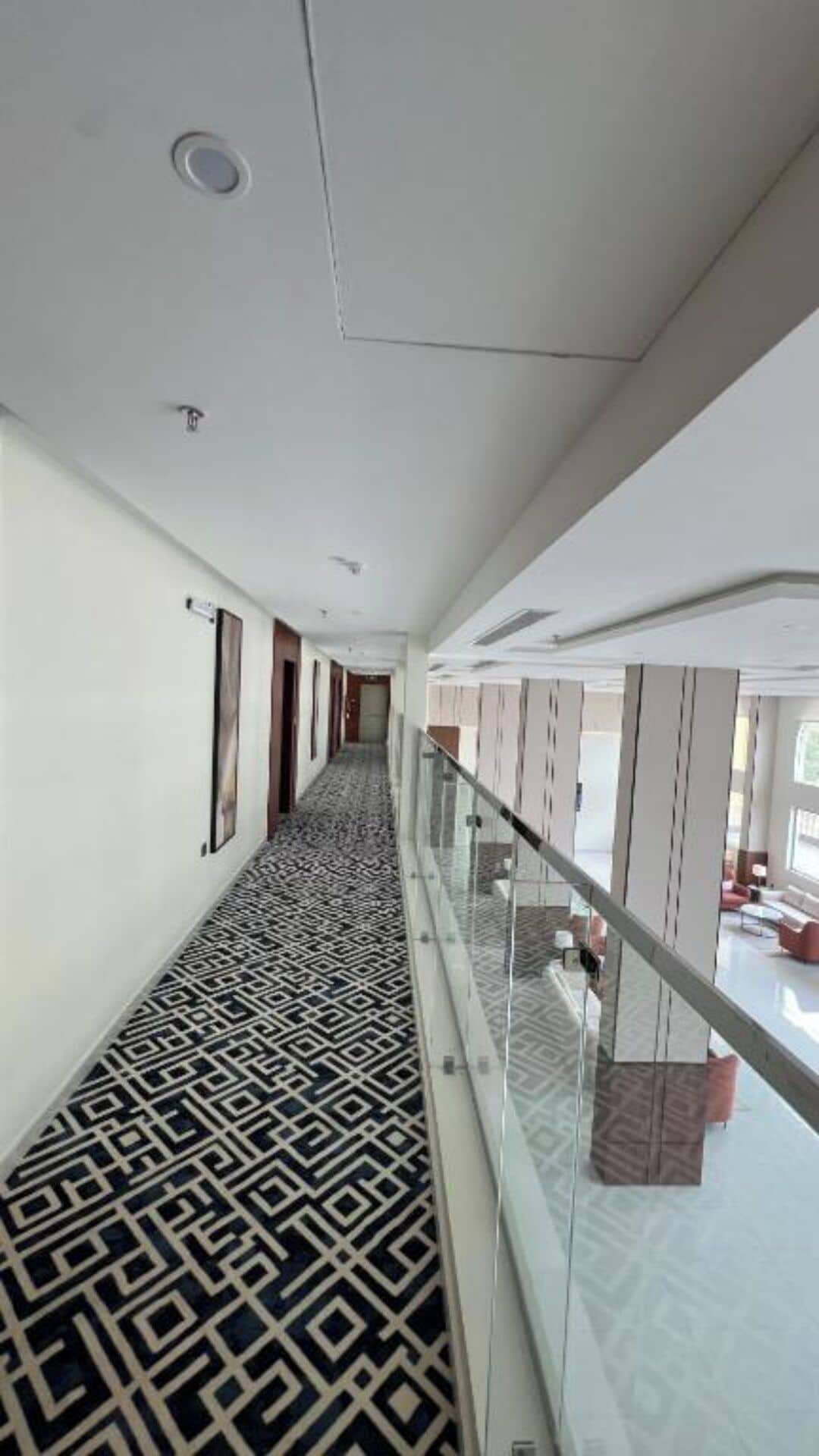 A corridor with patterned flooring, a glass railing, and a view of a room with furniture.