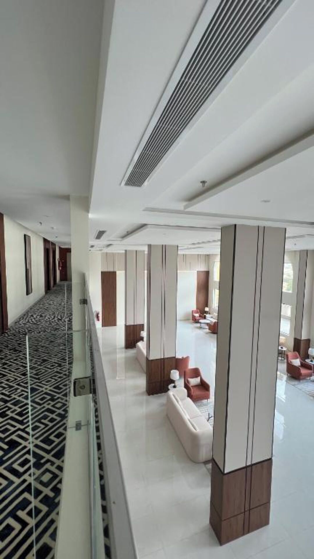 A modern hotel lobby with a central seating area, a patterned carpet, and a glass railing overlooking the space.