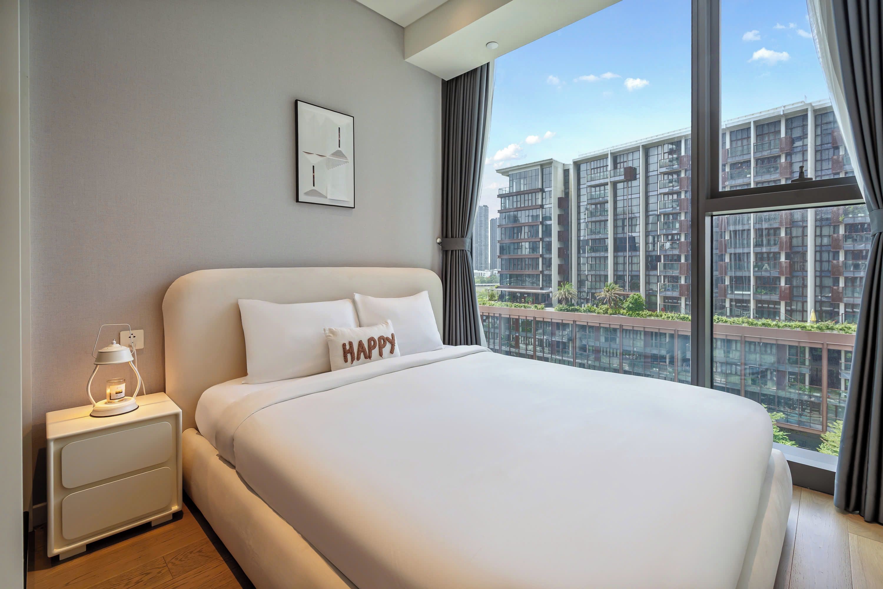 Une chambre d’hôtel moderne avec un grand lit, une table de chevet avec une lampe, un tableau encadré au mur et une vue sur les gratte-ciel par la fenêtre.