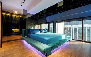 A modern bedroom with a large bed, a seating area, and a city view. - Metropole Riverside Luxury Apartment (Thu Duc)