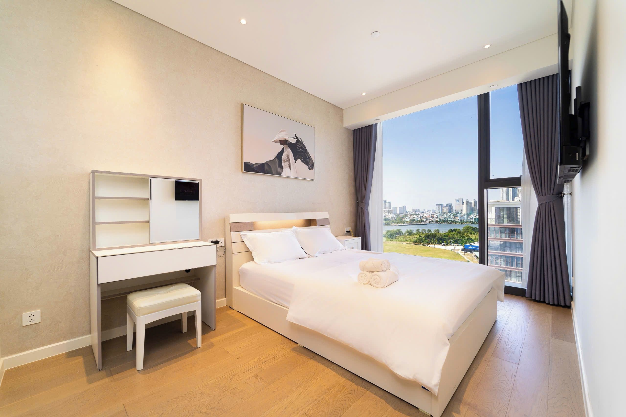 Habitación de hotel con cama, escritorio, silla, ventana grande con vista a la ciudad y un cuadro en la pared.