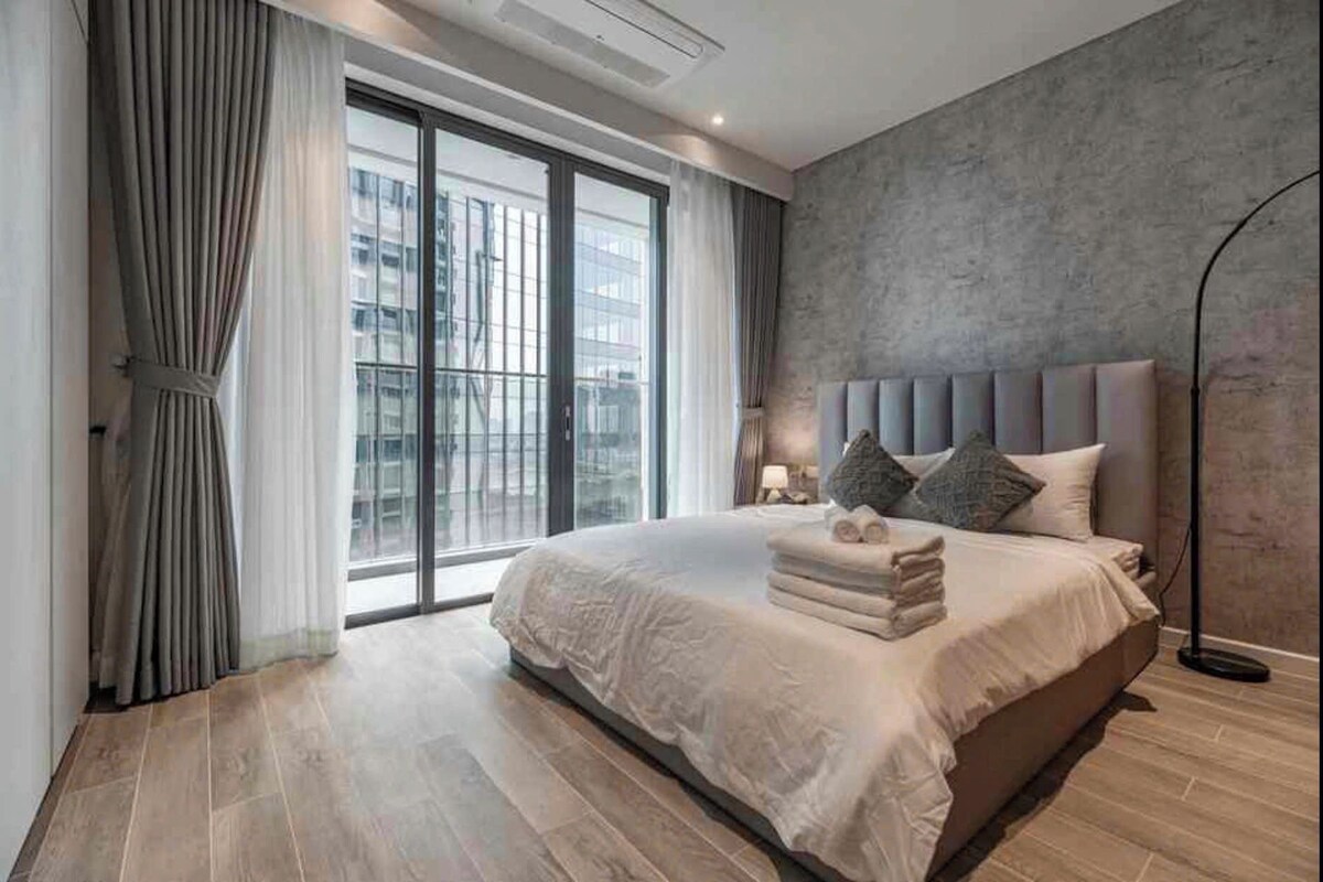 A modern bedroom with a large bed, a city view, and a concrete accent wall.