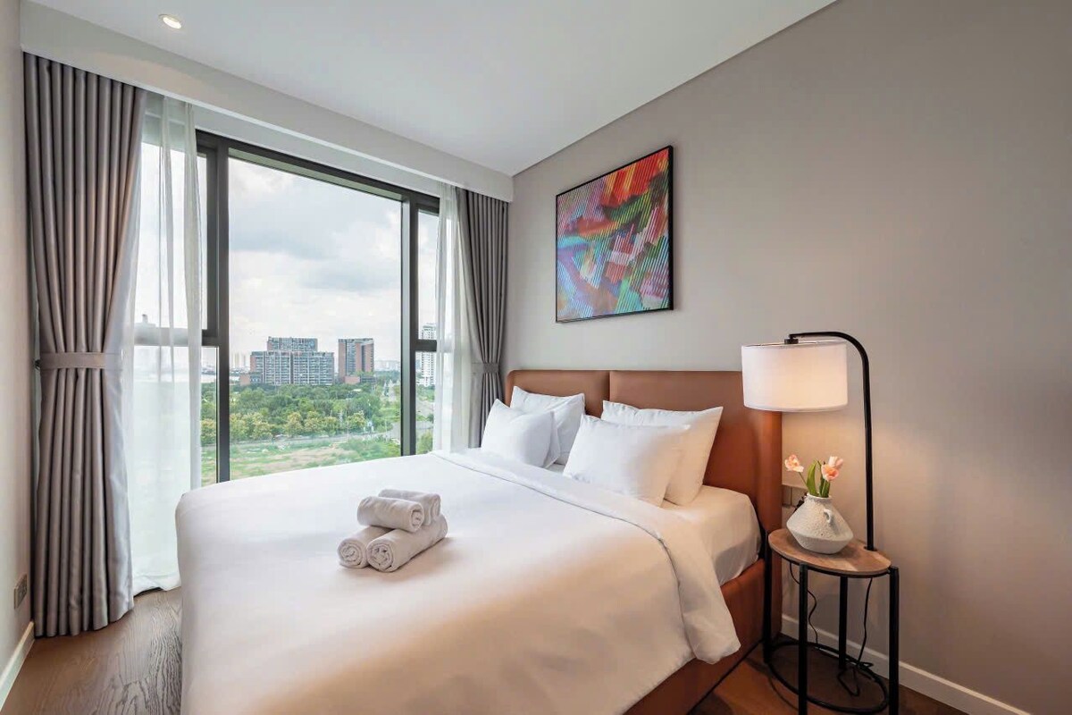 A hotel room with a large bed, a bedside table with a lamp, and a city view through the window.