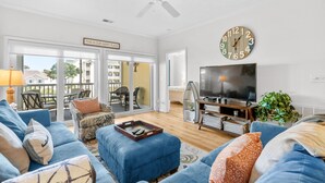 A living room with a blue sofa, a TV, a clock, and a ceiling fan. - 10% off 3-7 nights thru Oct. 2BR 2BA condo. 3rd flr. Elev. Pool view. Short walk to beach. (North Myrtle Beach)