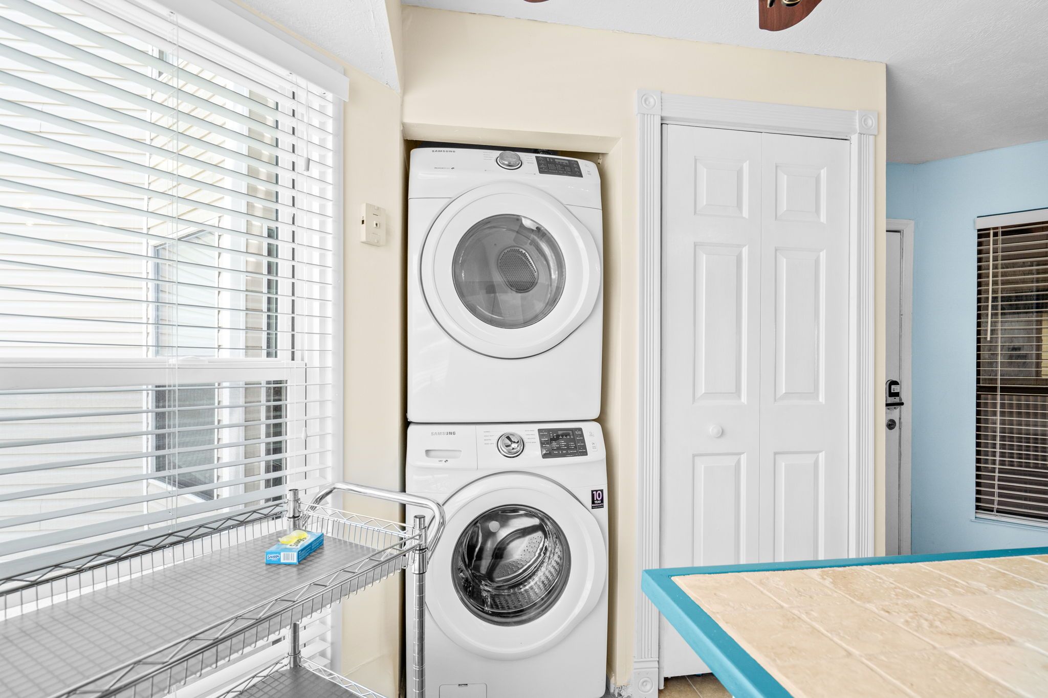Une buanderie équipée de deux machines à laver et deux sèche-linge superposés, d’un étagère métallique contenant des produits lessiviels et d’une porte située sur le côté droit.