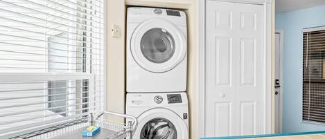 Une buanderie équipée de deux machines à laver et deux sèche-linge superposés, d’un étagère métallique contenant des produits lessiviels et d’une porte située sur le côté droit.