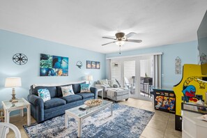 A living room with a blue sofa, a coffee table, and a ceiling fan. - Beachside Bliss. 2nd row Windy Hill. Outdoor pool. Perfect location! (North Myrtle Beach)