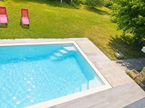 A rectangular swimming pool with clear blue water, surrounded by a tiled deck and a grassy lawn with two red lounge chairs.