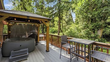 Una terraza de madera con jacuzzi, mobiliario exterior y una zona cubierta con luces colgantes.