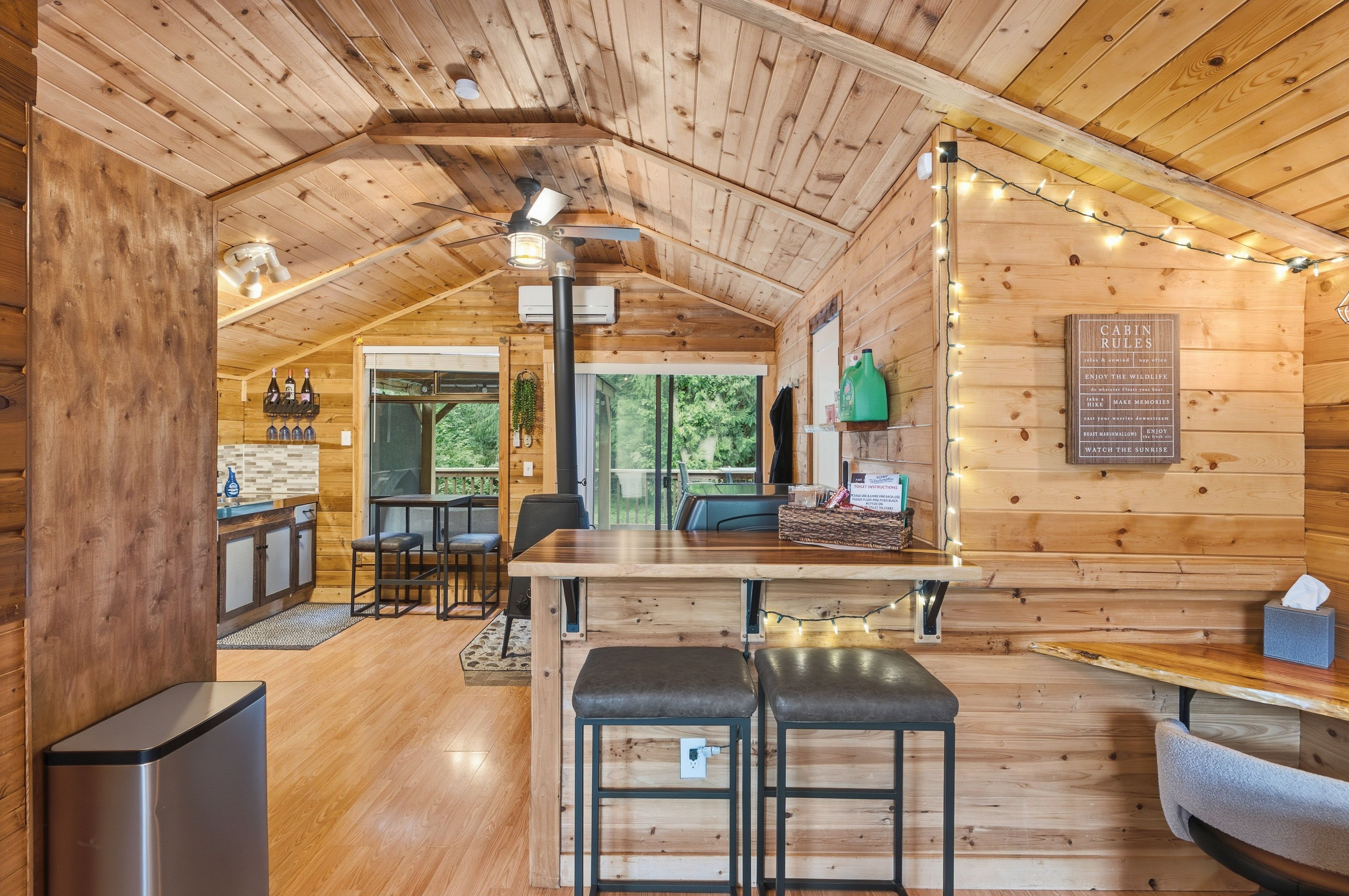 A cozy cabin interior with a wooden ceiling, a dining table with two stools, a kitchen area with a sink, and a large window.