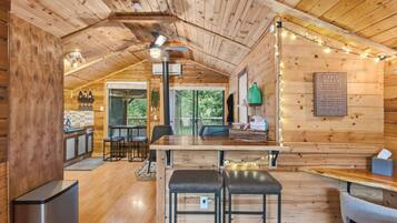 A cozy cabin interior with a wooden ceiling, a dining table with two stools, a kitchen area with a sink, and a large window.