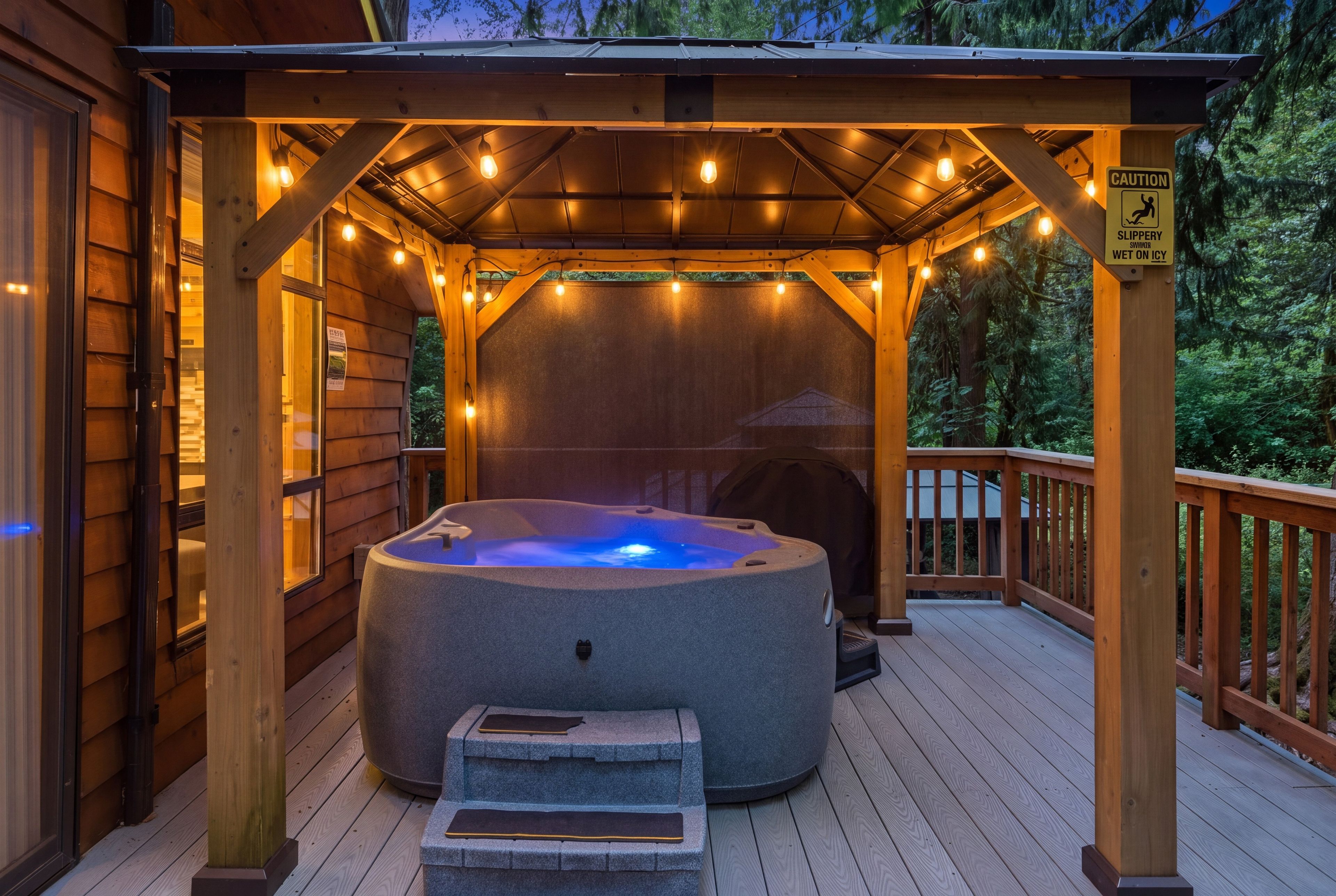 Un jacuzzi bajo un porche de madera con luces colgantes, rodeado por una terraza de madera y un paisaje boscoso.