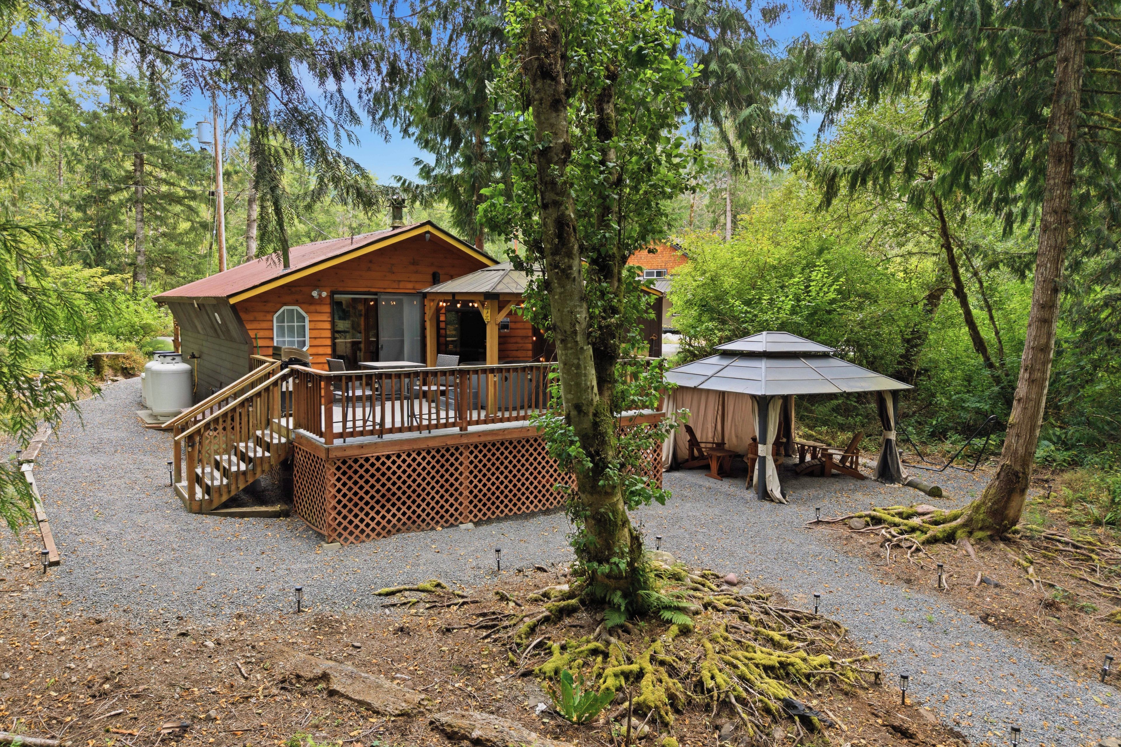 A cabin with a deck, a covered outdoor seating area, and a gravel driveway surrounded by trees.