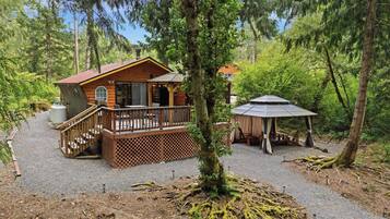 A cabin with a deck, a covered outdoor seating area, and a gravel driveway surrounded by trees.