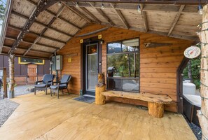 Uma cabana de madeira com varanda coberta, duas cadeiras, um banco e uma janela grande.