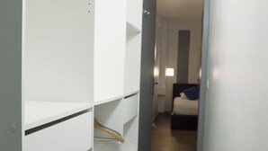 Un couloir étroit, avec des étagères blanches d’un côté, et une chambre au bout, où se trouve un lit recouvert d’une couverture bleue.