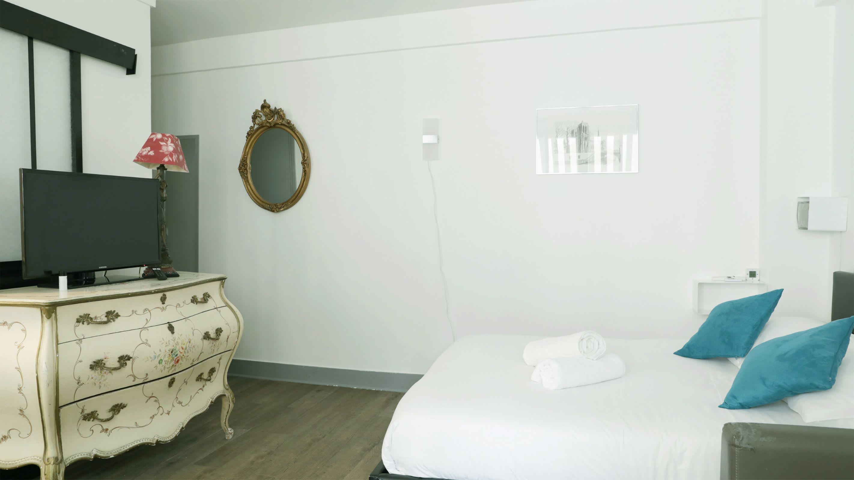Hotellihuone, jossa on sänky, lipasto, televisio, peili ja ikkuna, jossa on sälekaihtimet.