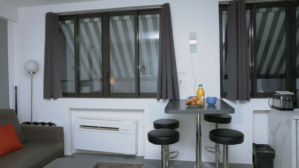 Un espace de vie compact comprenant un canapé, une petite table à manger avec des tabourets, un radiateur et une fenêtre avec des rideaux.