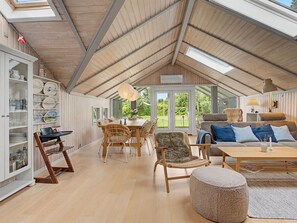 A spacious loft with a high ceiling, wooden beams, a dining area with wicker furniture, a cozy seating area with a sofa and ottoman, and a large window offering a view of the outdoors. - "Aleksis" - 900m from the sea by Interhome (Væggerløse)