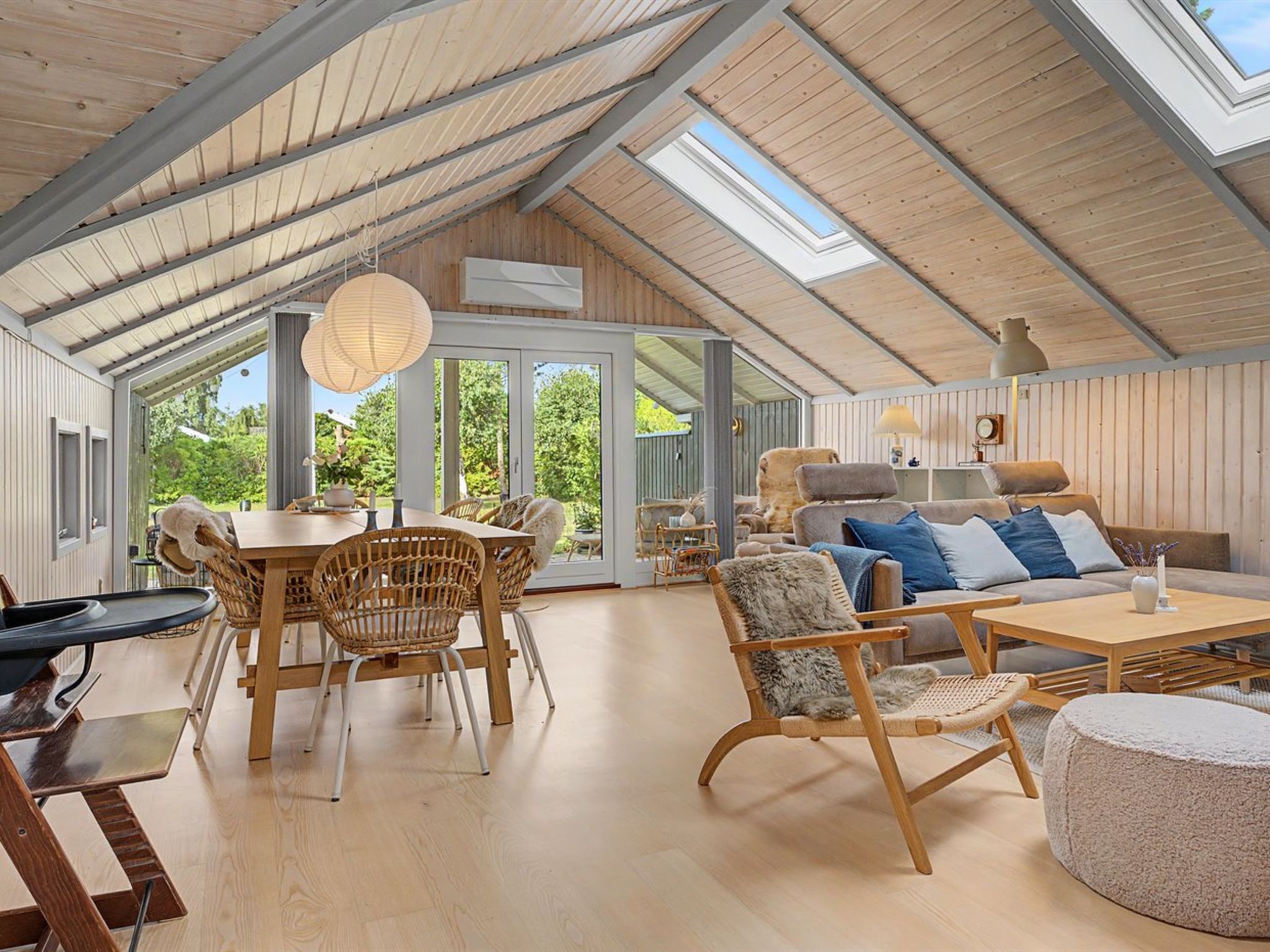 A spacious room with a high ceiling, wooden beams, a dining area with wicker chairs, a sofa set, and a large window offering a view of greenery.