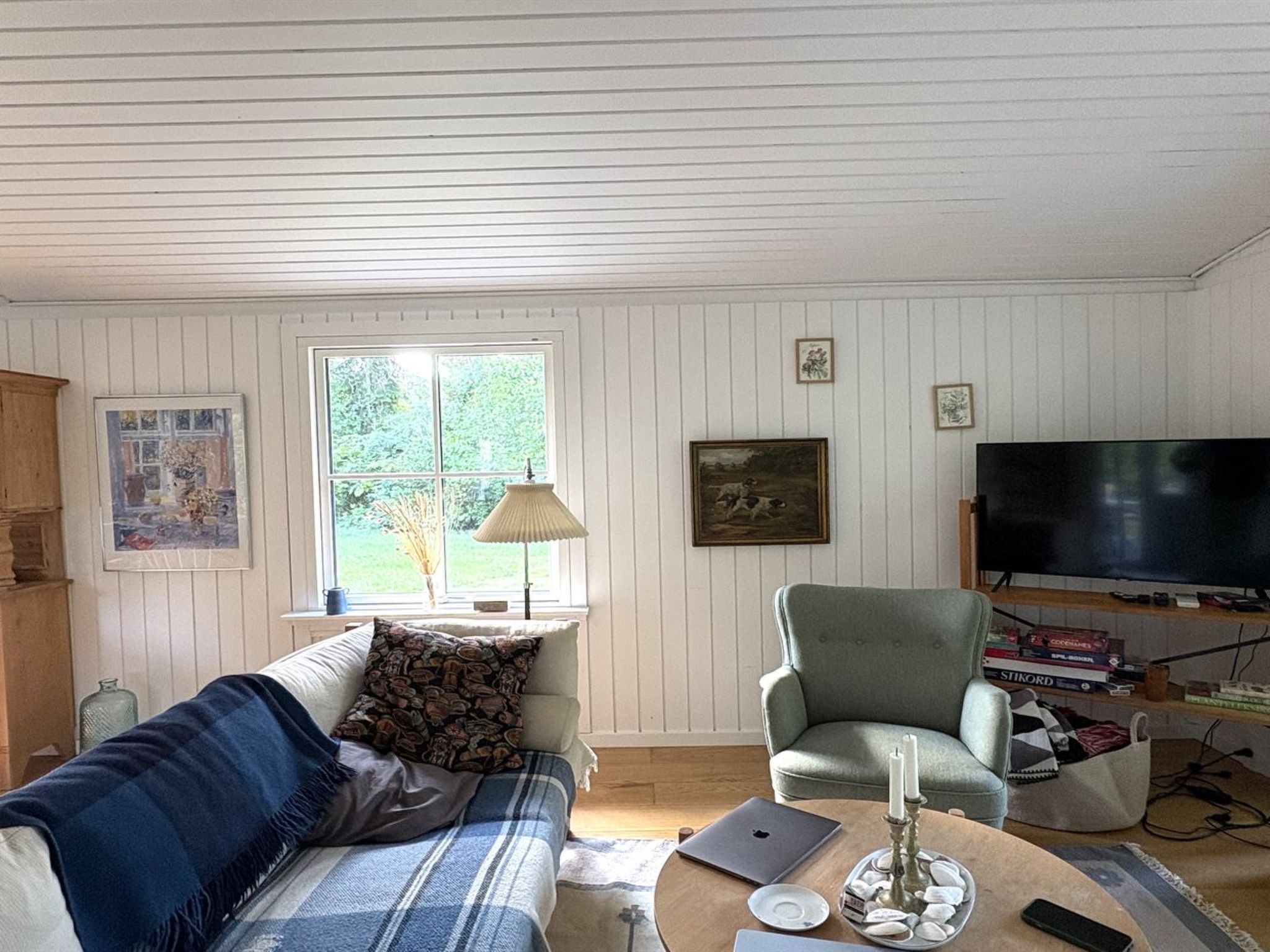 A living room with a sofa, armchair, and a television. There is a window with a view of greenery outside.
