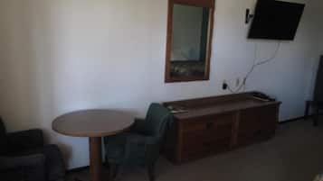 Ett rum med ett runt träbord, en grön fåtölj, en träbyrå, en platt-TV som är monterad på väggen och en spegel ovanför byrån.