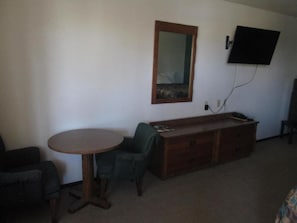 A room with a round wooden table, a green armchair, a wooden dresser, a flat-screen TV mounted on the wall, and a mirror above the dresser.