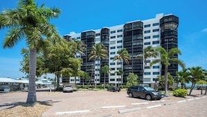 A multi-story hotel with a modern architectural design, featuring balconies and a parking area with cars and palm trees. - 2B/2B HARBOUR POINTE CONDO 715 ON THE BAY, FISHING DOCK, WALK TO BEACH! (Fort Myers Beach)