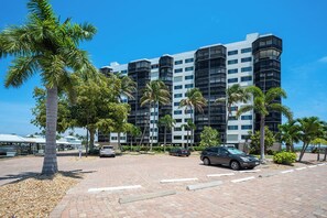 Un hotel de varios pisos con diseño arquitectónico moderno, que cuenta con balcones y un estacionamiento con coches y palmeras.