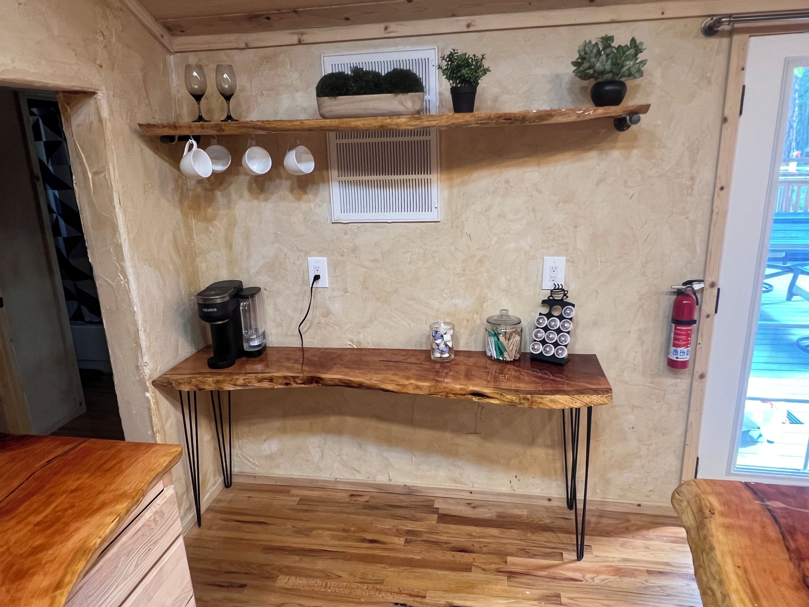 Une cuisine avec une table en bois, une machine à café, un extincteur et des étagères où sont rangés des verres et des plantes.