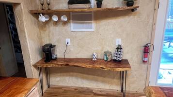 Une cuisine avec une table en bois, une machine à café, un extincteur et des étagères où sont rangés des verres et des plantes.