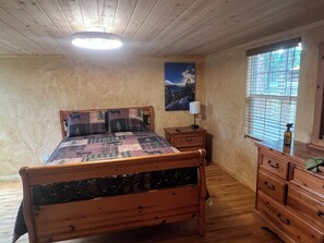 Una cama de madera con colcha, una mesita de noche con lámpara, un aparador y una ventana con persianas.