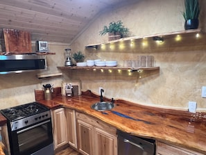 Una cocina con encimera de madera, estufa, horno y lavavajillas. Hay estantes con vajilla y una planta.